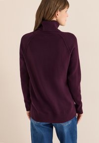 Maglione a collo alto di colore viola scuro con un punto a rilievo, colletto e orlo a coste, e maniche raglan. Indossato con jeans in denim blu.