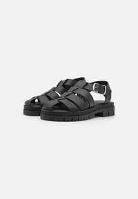 Sandalias negras con múltiples correas anchas, con los dedos al aire y una correa en el tobillo con hebilla. Suela de goma gruesa con un patrón de relieve texturizado.