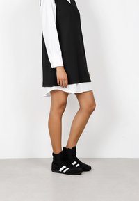 Abito nero senza maniche sovrapposto a una camicia bianca con bottoni, abbinato a sneaker alte nere con tre righe bianche.