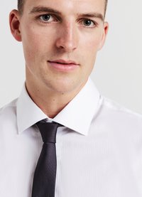 Jeune homme avec les cheveux courts portant une chemise blanche et une cravate noire texturée, sur un fond clair uniforme.