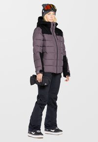 Lilla og sort puffy jakke med lynlås foran, sorte skibukser og sorte handsker. En stripet hue med skibriller fuldender vinteroutfittet.