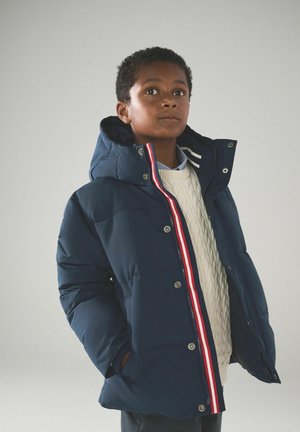 Giubbotto imbottito blu navy con cappuccio, dotato di strisce decorative rosse e bianche, bottoni a pressione argentati e tasche laterali. Indossato sopra un maglione crema.