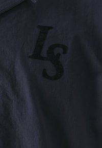 Tummansininen takki, jossa on teksturoitu kangas, jossa on pystysuorat raidat, ja mustasta samettimateriaalista valmistettu "LS"-logo edessä.