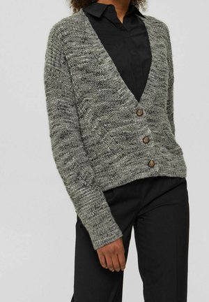 Une femme porte un cardigan gris texturé boutonné par-dessus une chemise noire à col et un pantalon noir, avec sa main gauche détendue le long du corps.