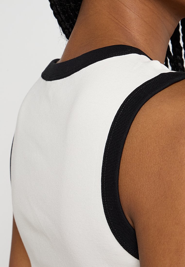 Biały top bez rękawów z czarną prążkowaną dekoltem. Tkanina wydaje się gładka, a projekt posiada bezrękawowe otwory na ramiona, co podkreśla prosty styl.