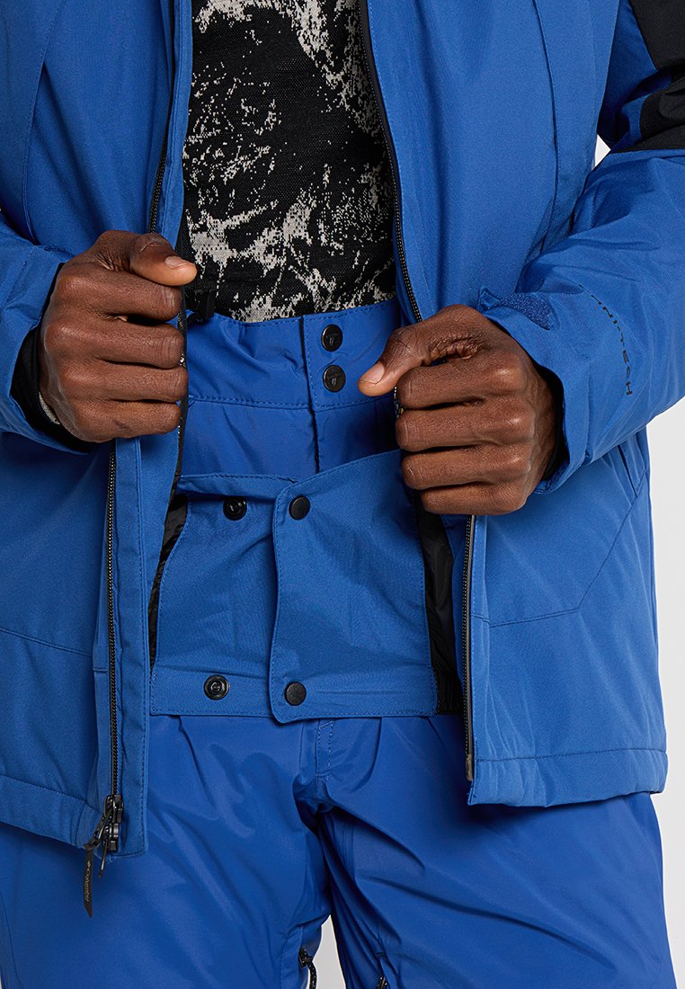 Hände, die eine blaue Skijacke und Hose mit mehreren Knöpfen und Reißverschlüssen über ein schwarz-weiß gemustertes Hemd anpassen.