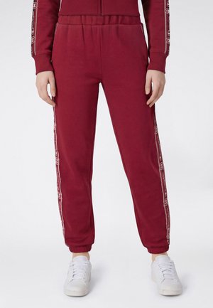 Nedre del af person iført vinrøde joggingbukser med sidestriber med logo og hvide sneakers på en ensfarvet lys baggrund.