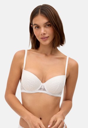 Mujer joven con cabello castaño hasta los hombros, vistiendo un sujetador balconette de encaje blanco, posando contra un fondo claro y liso.