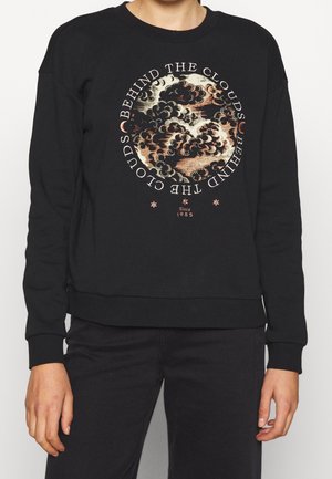 Personne portant un sweat-shirt noir avec un motif circulaire de nuage et le texte "Behind the clouds" et "Since 1985" sur le devant.