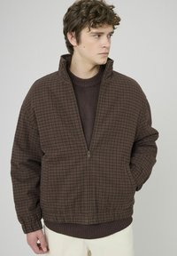 Veste à carreaux marron avec col montant, fermeture éclair à l'avant et ourlet côtelé. Texture douce, coupe oversize, portée sur un pull foncé.