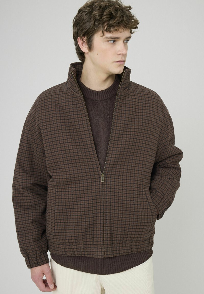 Veste à carreaux marron avec col montant, fermeture éclair à l'avant et ourlet côtelé. Texture douce, coupe oversize, portée sur un pull foncé.