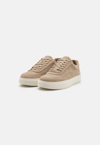 Beige suede sneakers with a rounded toe, flat laces, and a white rubber sole. Features stitching details and a low-profile design.