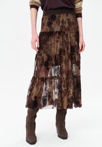 Bruine bloemenrok met meerdere lagen en een doorschijnende overlay, voorzien van donkere bloemenpatronen en een elastische tailleband. Gestyled met kniehoge bruine laarzen.