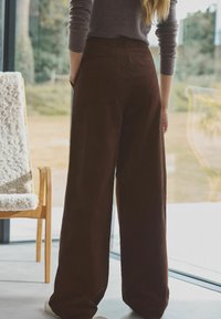 Brune brede corduroy bukser med høj talje, der har sidelommer og et blødt, tekstureret stof. Bagdetaljen viser et simpelt design.
