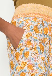 Calças com padrão floral e cintura elástica amarela; apresentam flores laranja e azuis sobre um fundo branco, e bolsos laterais.