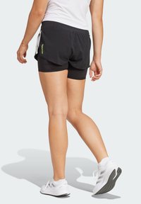Shorts de sport noirs avec un design superposé, ornés d'un logo vert fluo et de bandes blanches sur les côtés, associés à des chaussures de sport blanches.