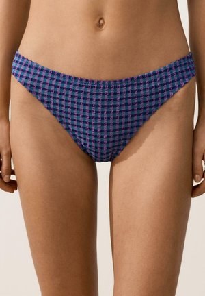 Žena v spodnej časti plaviek so vzorom fialovo-modrej kocky, stojaca s rukami uvoľnene pozdĺž tela, viditeľný stred tela a horné partie nôh.