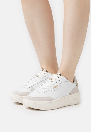 Sneaker bianca con accenti in suede, occhielli dorati e suola texture. Presenta un design low-top e materiali a contrasto. Logo del marchio visibile.