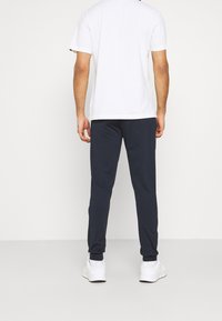 Mann steht mit dem Rücken zur Kamera, trägt ein weißes kurzärmliges Hemd, dunkle marineblaue Jogginghosen und weiße Sneakers vor einem schlichten Hintergrund.