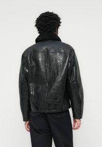 Veste en cuir noir avec une finition brillante, épaules larges et un col en fausse fourrure texturée noire. Présente une coupe ajustée et des coutures minimales.