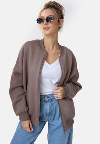 Brązowy, oversize'owy sweter z okrągłym dekoltem i ściągaczami, w połączeniu z białą koszulką z dekoltem w serek oraz niebieskimi jeansami o wysokim stanie.