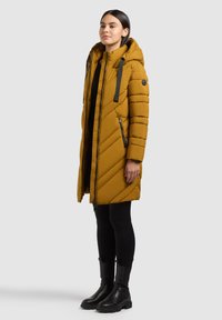 Femme debout portant un long manteau matelassé jaune moutarde à capuche, des leggings noirs et des bottines noires sur un fond gris uni.