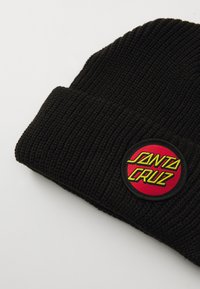 Beanie lavorato a maglia nera con una toppa rossa circolare che riporta la scritta "Santa Cruz" in giallo, caratterizzato da una texture a coste e un'ottima vestibilità.