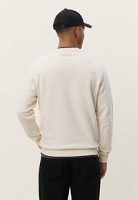 Sweat-shirt couleur crème avec un col rond, des poignets et un ourlet côtelés. Porté avec un pantalon foncé et une casquette bleu marine. Tissu lisse et doux, sans motifs visibles.