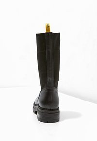 Svarta högstövlar med en slät läder tåhuvud, elastisk tygskaft och en gul dragflik med nummer 146 på baksidan.