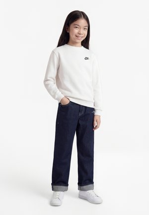 Κορίτσι που χαμογελά στέκεται με το ένα χέρι στην τσέπη, φορώντας λευκό φούτερ Nike, σκούρο τζιν με γυρισμένα μπατζάκια και λευκά αθλητικά παπούτσια σε λευκό φόντο.