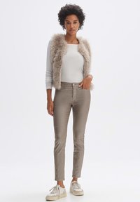 Beige Kunstfellweste über einem hellen Langarmshirt, kombiniert mit taillierten beige Hosen und weißen Sneakers. Einfaches Design, glatte Textur.