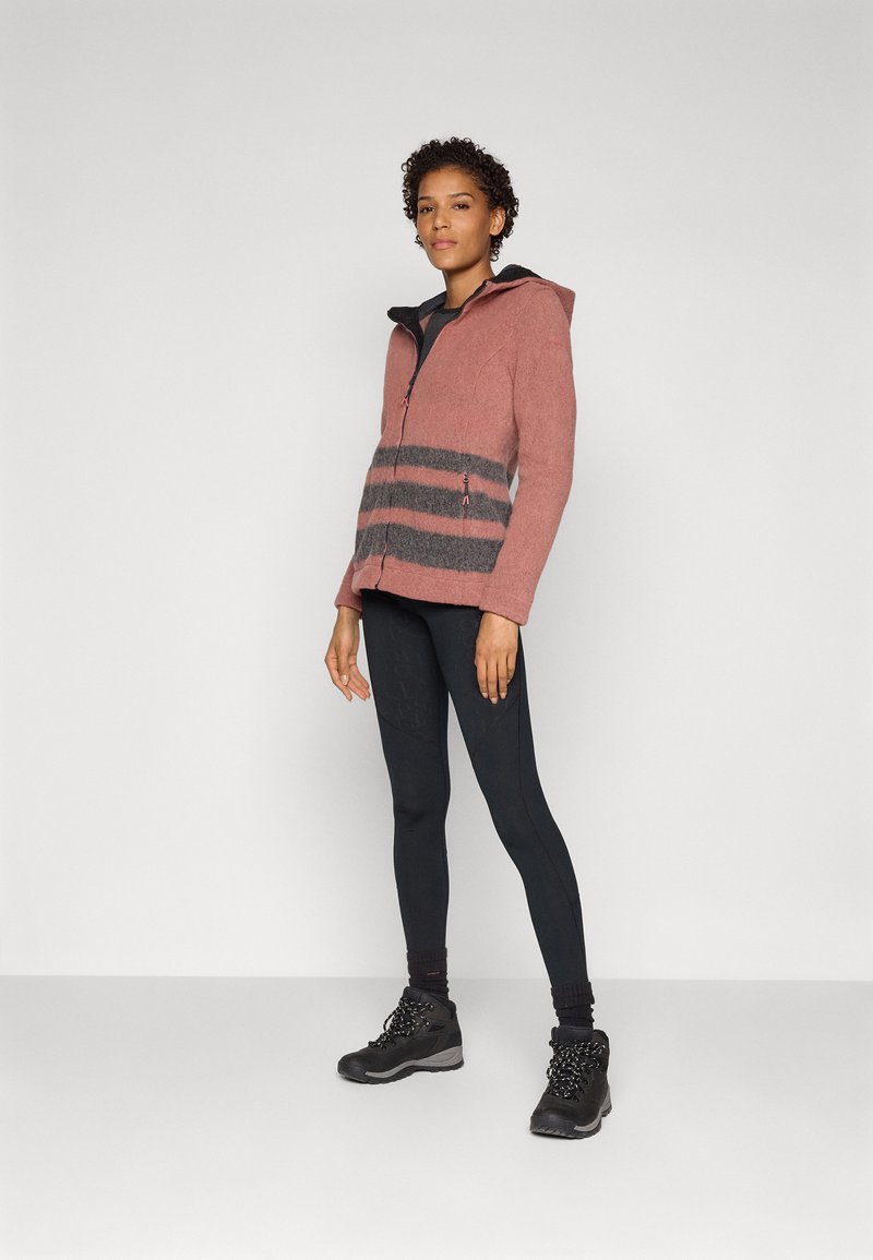 Donna in piedi con leggings neri, scarpe da trekking nere e una giacca con cappuccio rosa con zip e strisce grigie nella parte bassa anteriore.