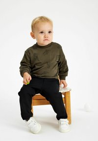 Sudadera verde oliva con estampados de dinosaurios negros, cuello de canalé; pantalones negros; zapatillas blancas; niño sentado en una silla de madera.
