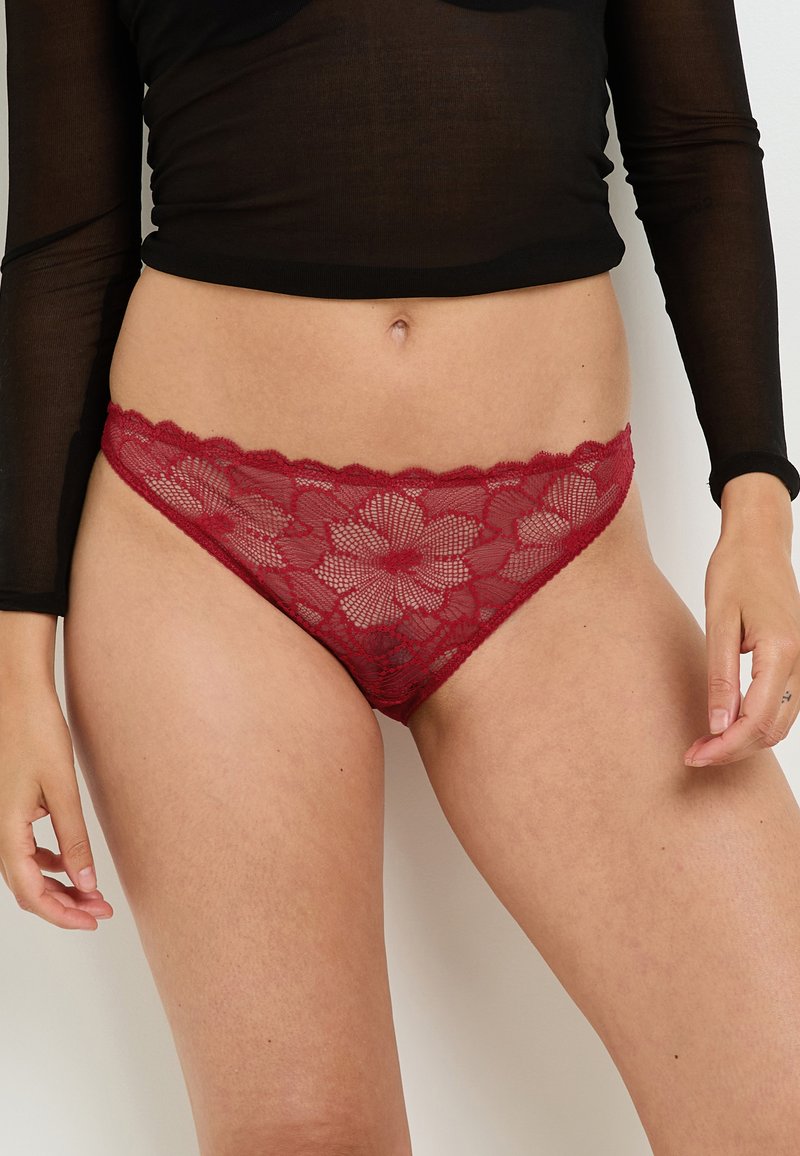 Tanga in pizzo rosso con motivi floreali, bordi scallop e una texture morbida. Indossato con un top nero trasparente a maniche lunghe.
