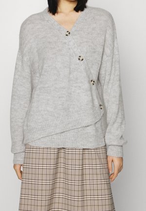Femme portant un pull en maille gris clair avec un détail de boutons en diagonale et une jupe à carreaux beige, montrée du cou jusqu'au milieu de la cuisse.
