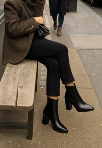 Bottes cheville en cuir noir avec un bout pointu et un talon épais, présentant une finition élégante et des accents dorés subtils.