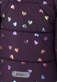 Veste violette gonflante avec un motif de cœur métallique irisé. Dispose d'un patch logo visible et d'une texture matelassée.