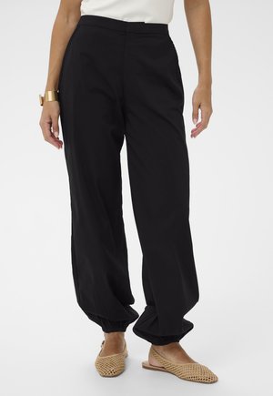 Femeie purtând pantaloni negri largi cu mansete elastice si balerini bej cu bareta, stand in picioare pe un fundal alb simplu.