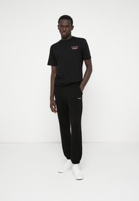 Fekete pamut póló elöl logóval, fekete melegítőnadrággal kombinálva, amely elasztikus mandzsettákkal és oldalsó zsebbel rendelkezik. A fehér sportcipők teszik teljessé az öltözéket.