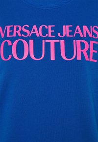Tissu bleu avec un grand texte rose indiquant « Versace Jeans Couture » sur la zone de la poitrine d'un vêtement.