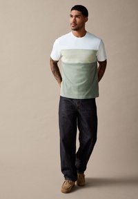 Rövid ujjú póló fehér felső résszel, világoszöld középső panellel és sötétebb zöld alsó résszel, sötét farmernadrággal és bézs lábbelivel kombinálva.