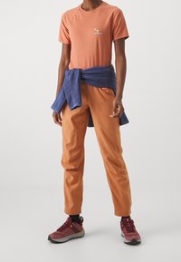 Camiseta naranja de manga corta con un pequeño logo, combinada con pantalones naranja y zapatillas deportivas color burdeos. Suéter azul atado a la cintura.