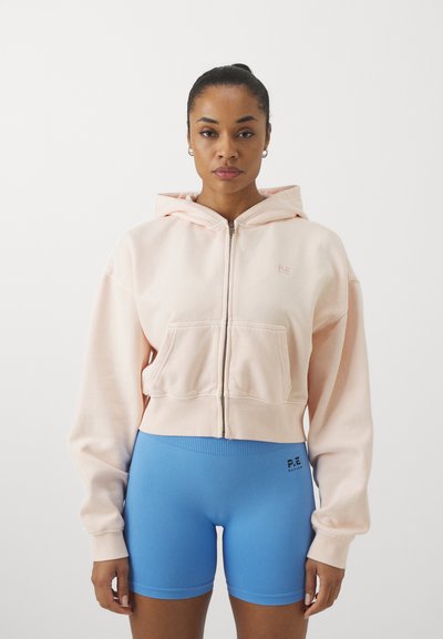 Hoodie court rose clair avec une fermeture éclair et une poche avant, associé à un short bleu ajusté arborant un logo. Texture de tissu lisse.