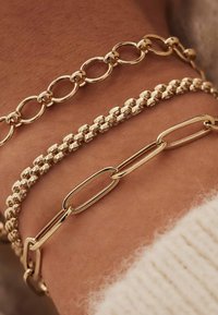 Drei goldene Armbänder in unterschiedlichen Designs: eine verkettete Kette, ein Perlenmuster und eine langgestreckte ovale Kette, alle mit einem glänzenden Finish.
