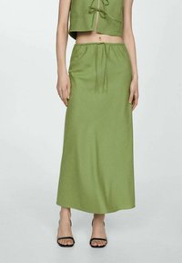 Frau in einem grünen hochgeschnittenen Midi-Rock mit Kordelzug und schwarzen offenen Absatzschuhen, die vor einem einfachen hellen Hintergrund steht.
