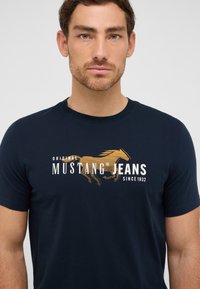 Tamsiai mėlyna medvilninė marškinėliai trumpomis rankovėmis, su auksine arklių grafika ir baltu tekstu "MUSTANG JEANS". Apvalios iškirptės dizainas.