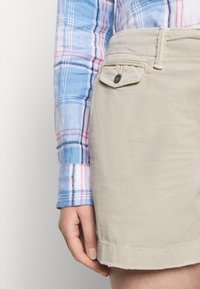 Osoba nosící modrou a růžovou kostkovanou košili zastrčenou do béžových kraťasů s kapsou na knoflík poblíž boků.