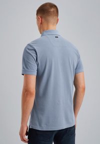 Homme portant un polo à manches courtes bleu clair et un jean foncé, debout de dos contre un fond gris uni.