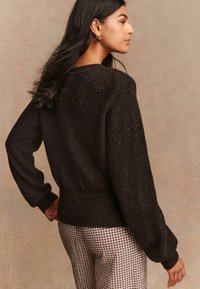 Übergroßer schwarzer Pullover mit strukturiertem Strick, feinen metallischen Punkten und geripptem Saum. Das Modell trägt karierte Hosen und zeigt die Rückansicht.