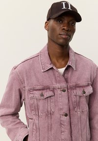 Φωτεινό ροζ τζιν μπουφάν με δύο τσέπες στο στήθος και κουμπιά, φορεμένο πάνω από λευκό πουκάμισο, συμπληρωμένο με καφέ καπέλο με λευκά γράμματα.